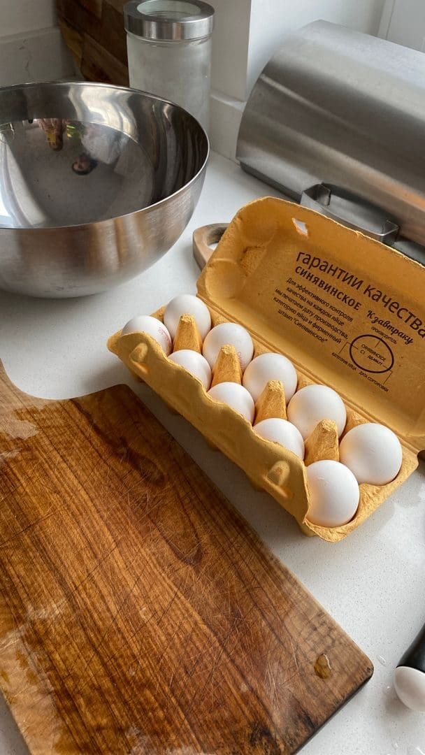 Open egg carton on a wooden chopping board with a stainless steel bowl, ideal kitchen preparation scene.
