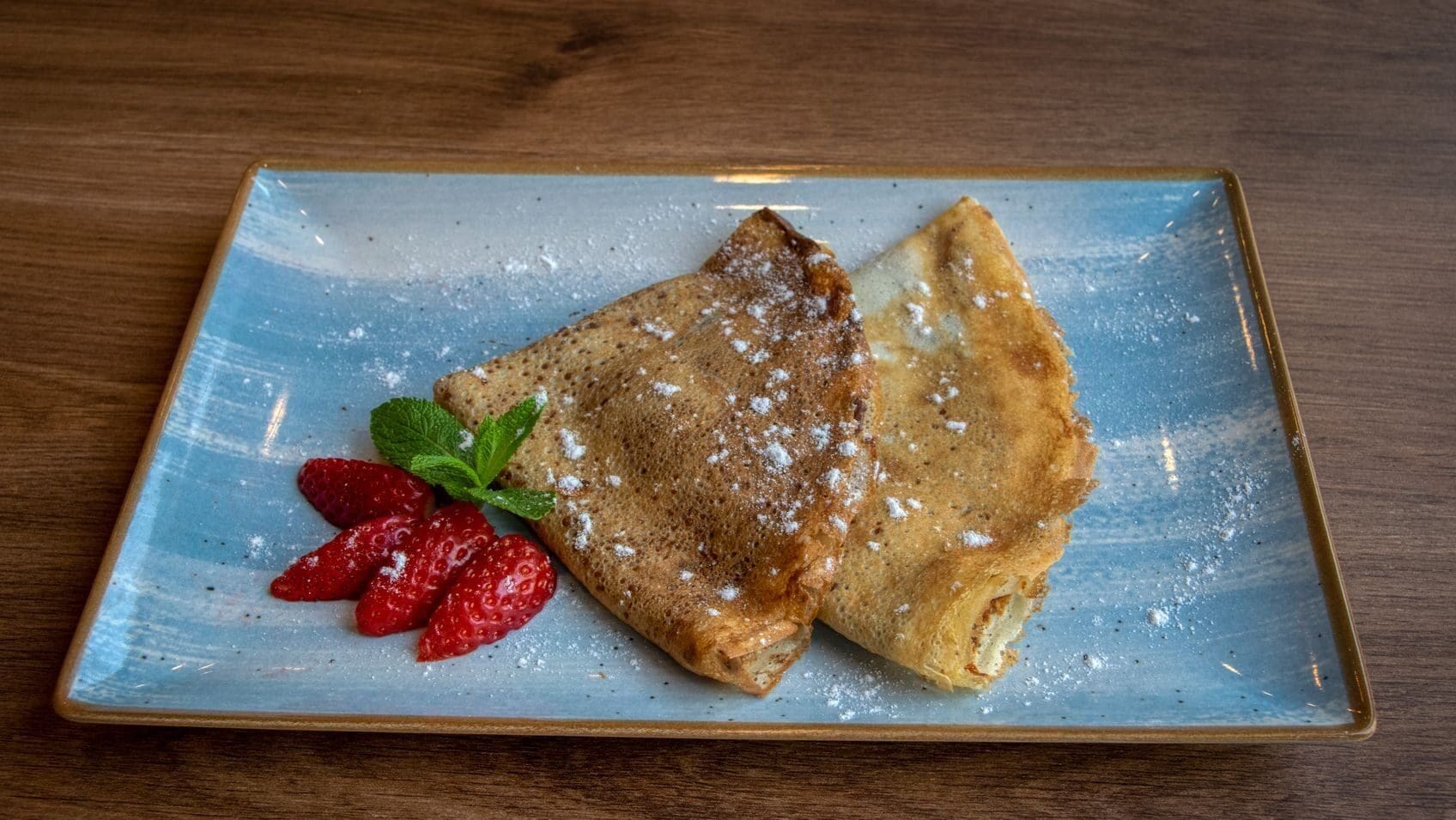 A plate of crêpes dusted with powdered sugar, served with strawberries and mint.