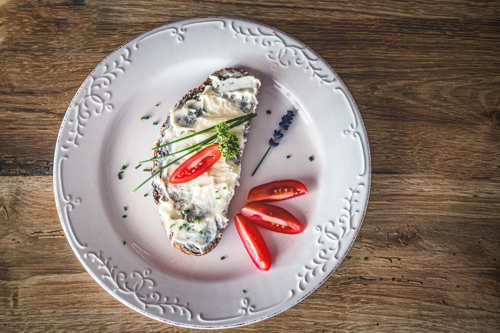 A rustic open-faced sandwich topped with creamy spread and garnished with cherry tomatoes, herbs on a decorative plate.