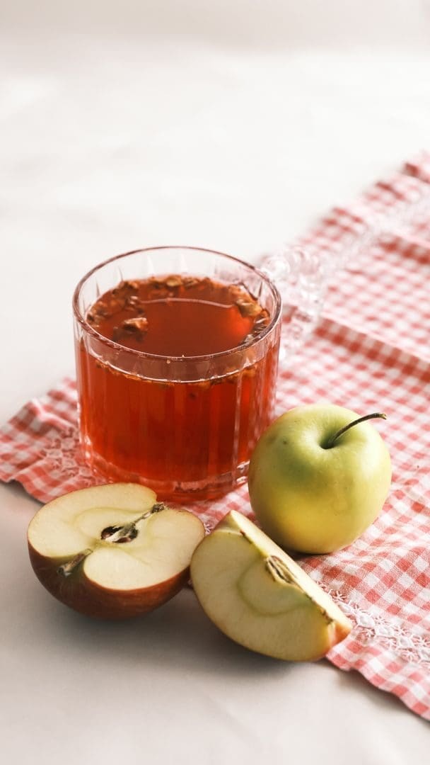 A clear glass of apple tea next to whole and sliced apples on a checkered cloth.