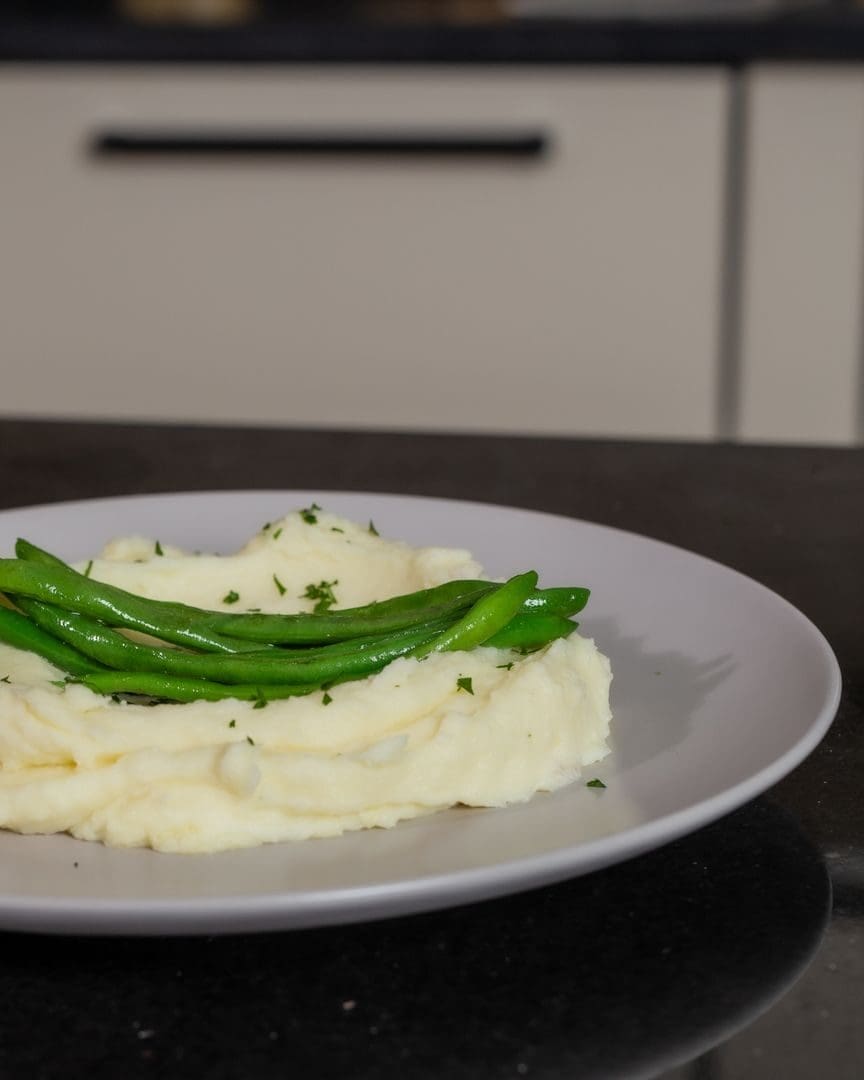 Plate of creamy mashed potatoes topped with fresh green beans, garnished with herbs.