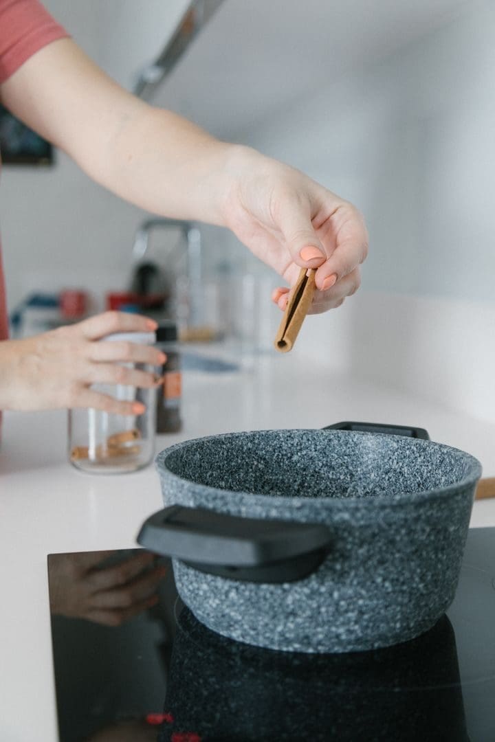 Close-up of hand adding cinnamon stick to a pot on a stovetop in a modern kitchen.