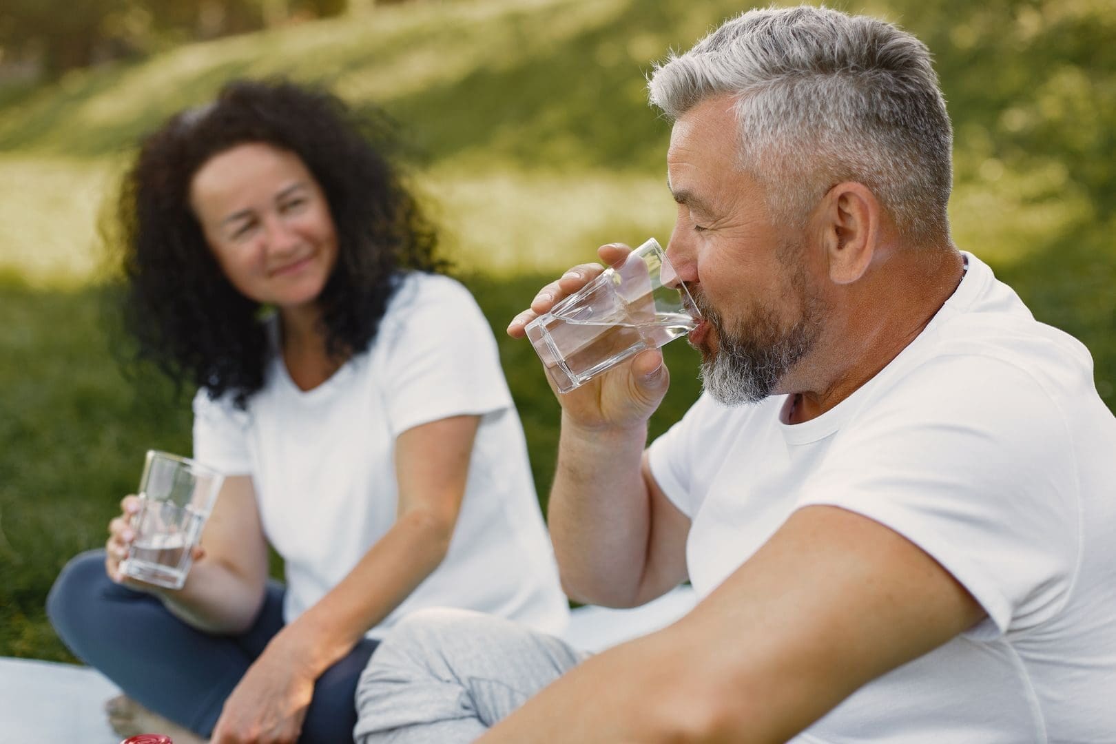 A senior man and woman sit outdoors, smiling while drinking water, enjoying a sunny day.