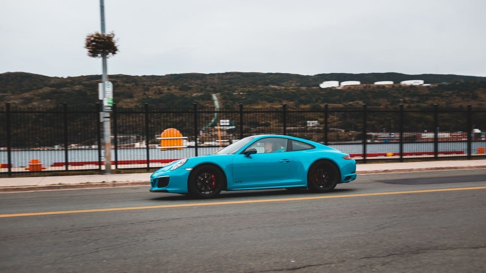 Side view of contemporary blue car driving on asphalt roadway near metal fence along embankment with sea with mountains in distance